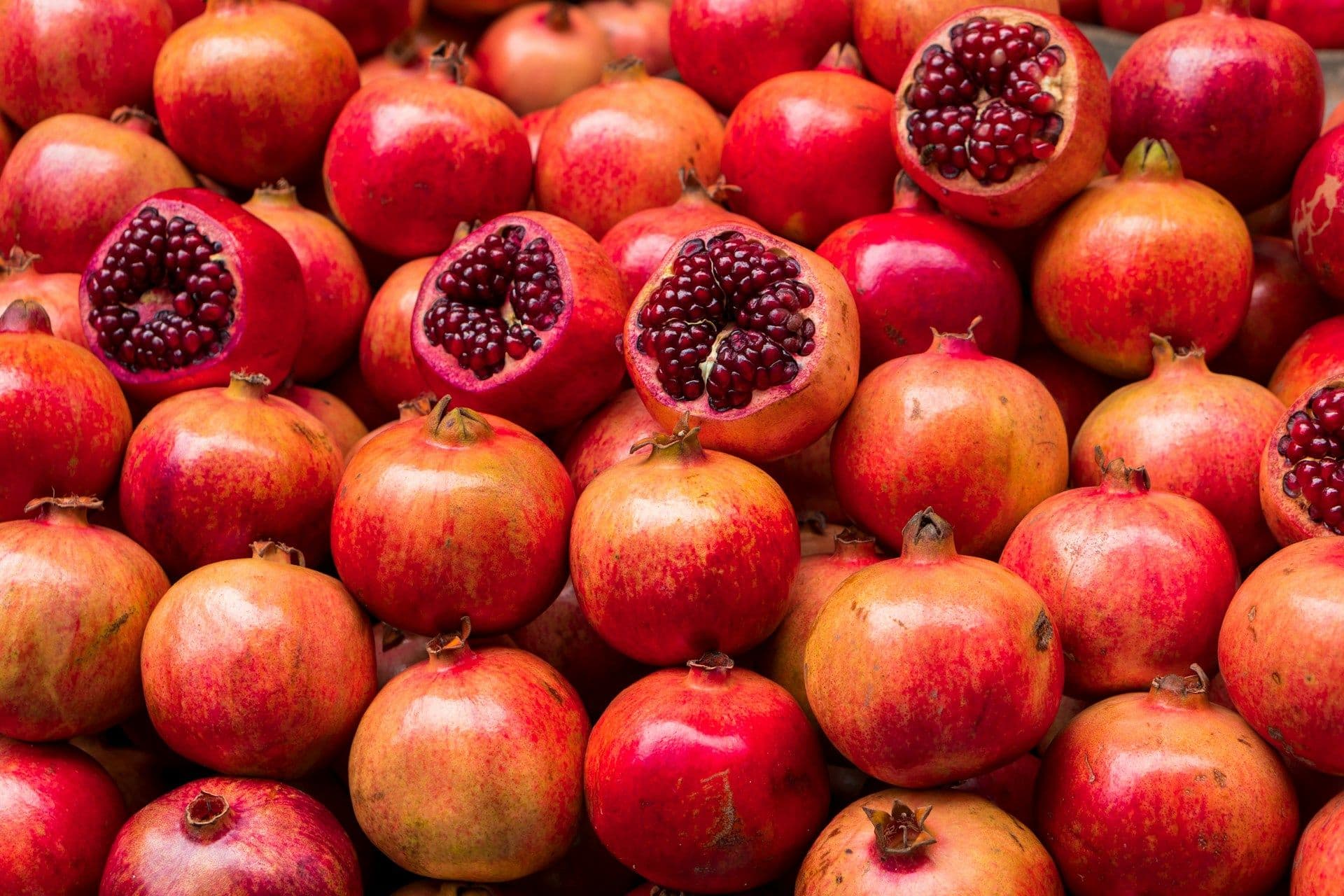 A large pile of fresh pomegranates, some of which are cut open to reveal their vibrant, red, juicy seeds.